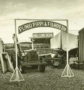 Caminhão antigo sob um arco com a inscrição Plínio Pippi & Filhos Ltda, Ind. Com. Repr.